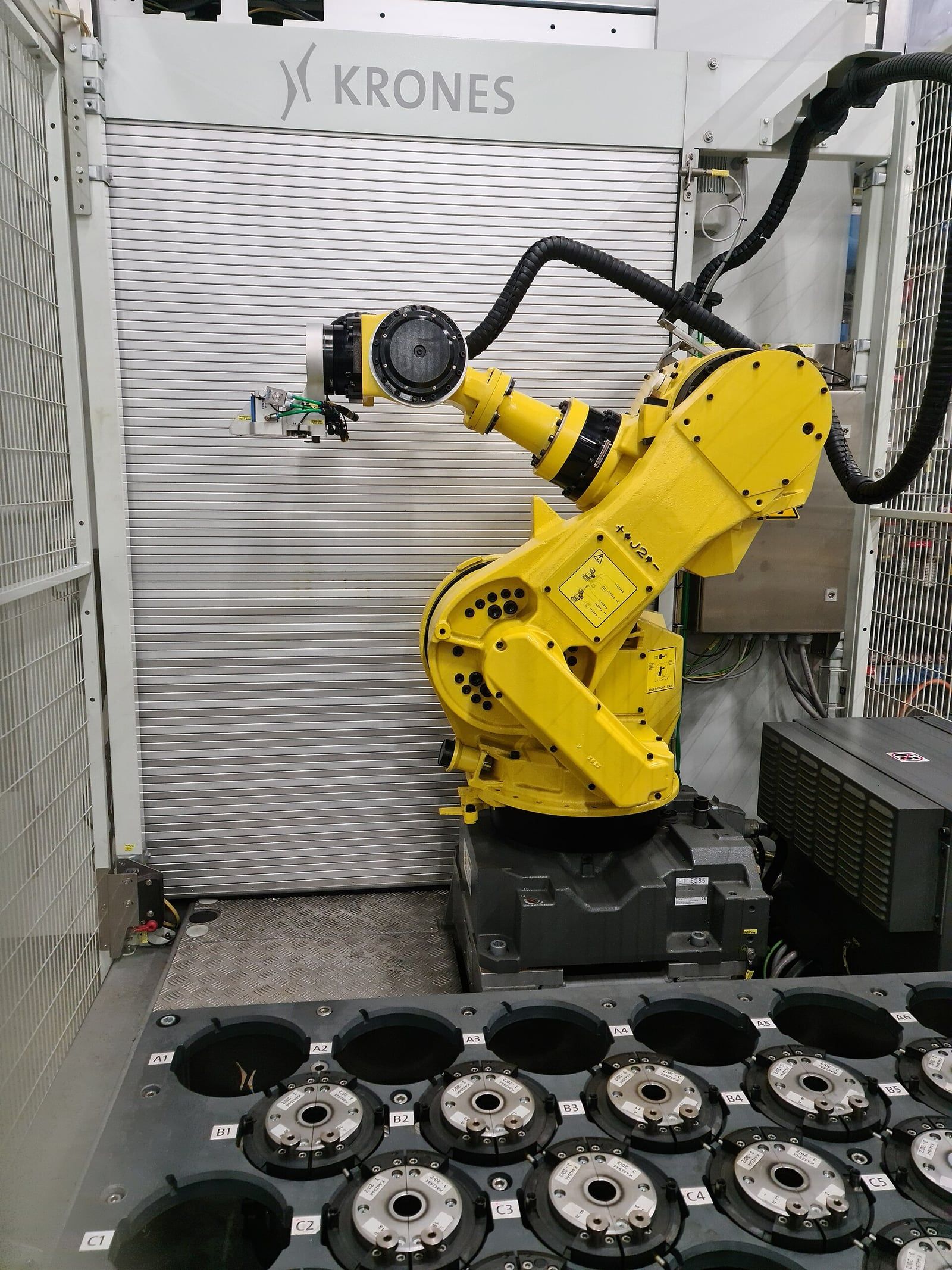 Bras robotisé industriel jaune Fanuc M-710iC installé dans une cellule grillagée, devant un rideau métallique portant l'inscription Krones, au-dessus d'un plateau de pièces numérotées.