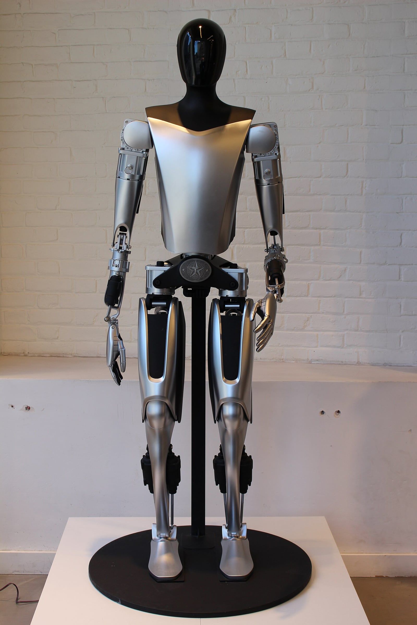 Robot humanoïde Tesla Optimus, d'une première génération, exposé debout sur un socle noir devant un mur de briques blanches. Il a une tête noire brillante, un torse argenté et des articulations mécaniques apparentes.